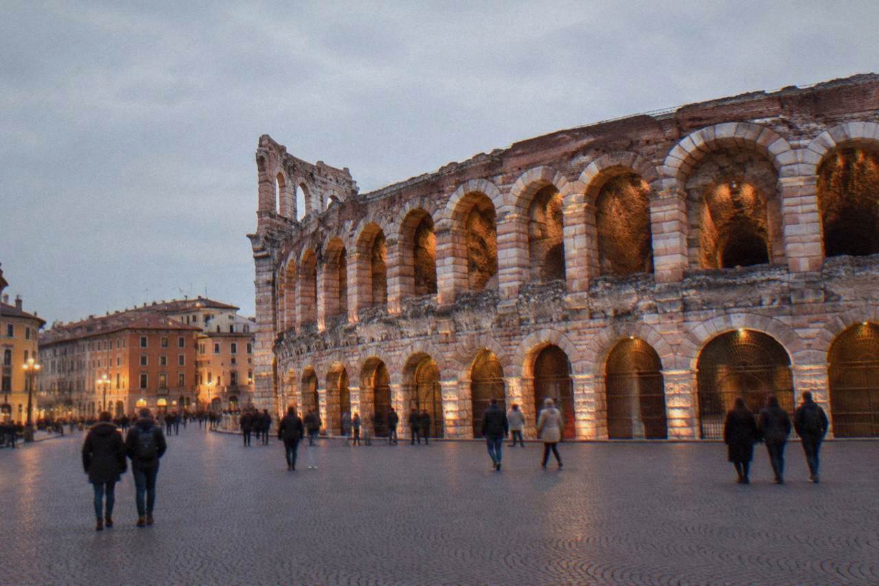 The floodlit Roman Arena of Verona at dusk seen from Piazza Bra, with evening visitors walking past the arches