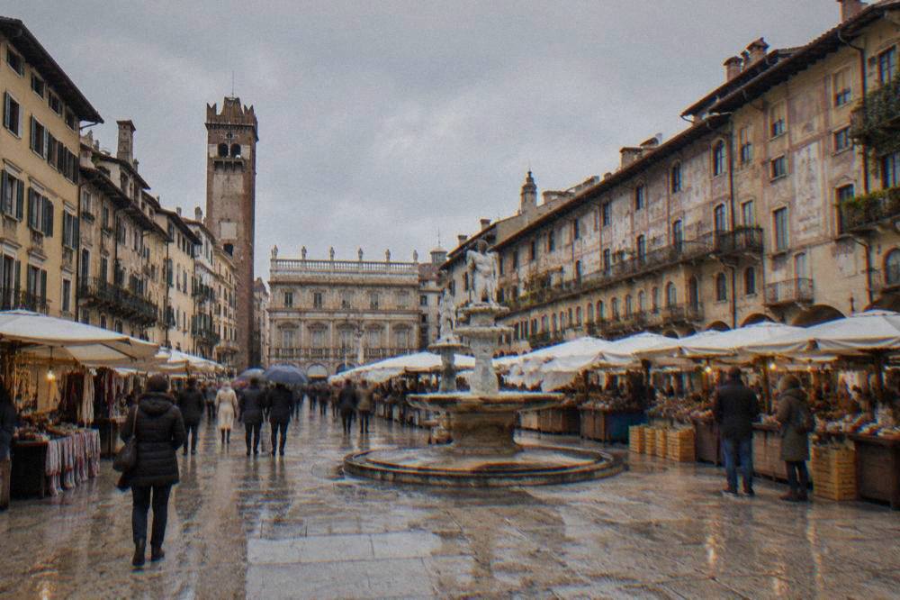 Visitors standing in the open square of Piazza delle Erbe in Verona among market stalls and old painted buildings