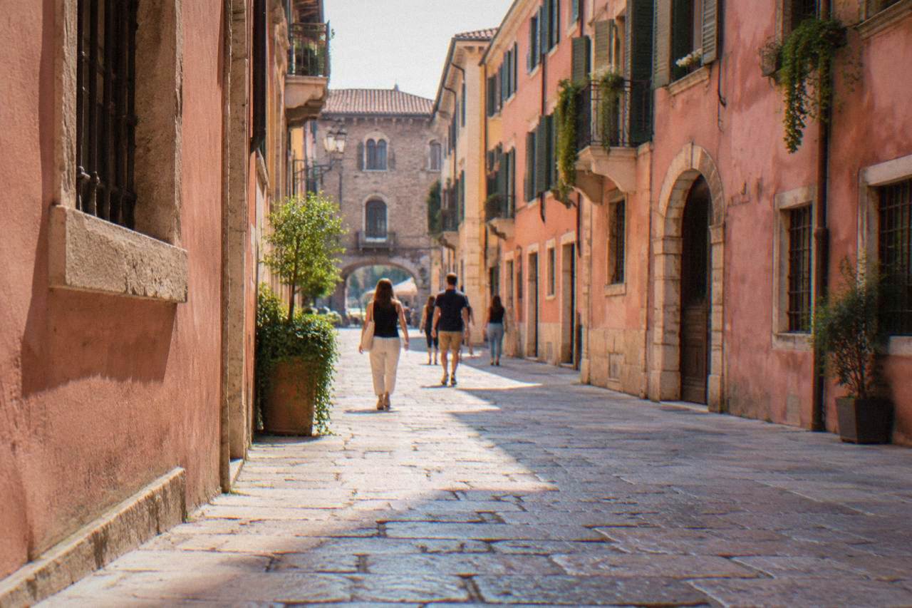 Pedestrians walking along a narrow cobbled street of old Verona with rose-pink buildings on both sides