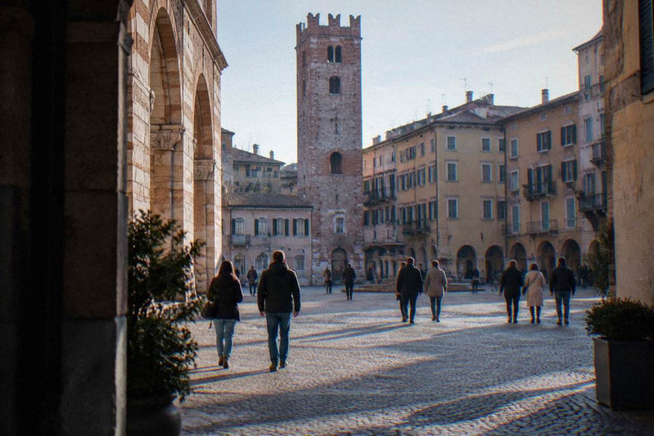 Visitors in light jackets walking across a cobblestone square in Verona with an old brick tower behind them