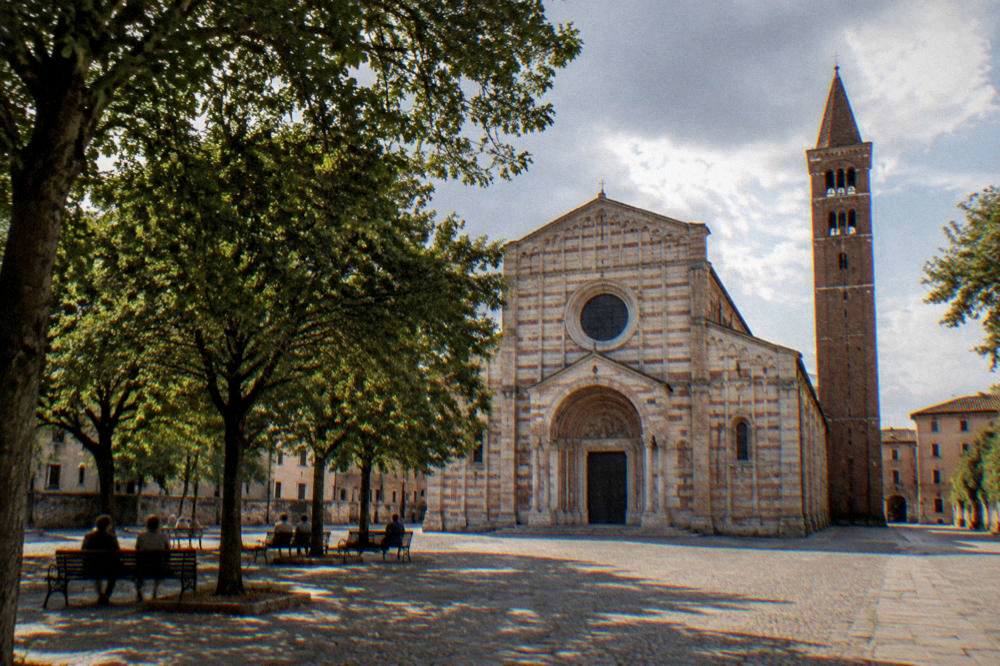 A broad square with trees in front of a large church, with people sitting on benches in the shade