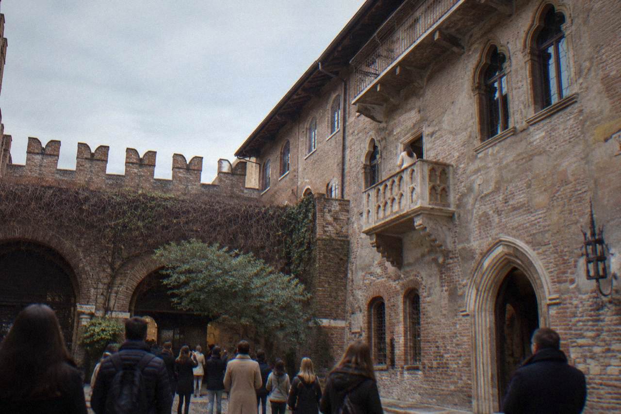A medieval brick courtyard in Verona with a stone balcony on an upper facade and visitors gathered below