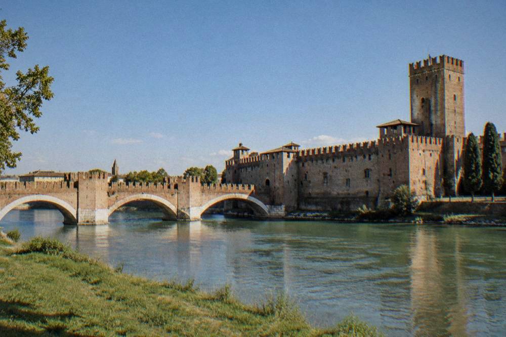 Bridge arches over the Adige seen from the grassy riverbank below the castle