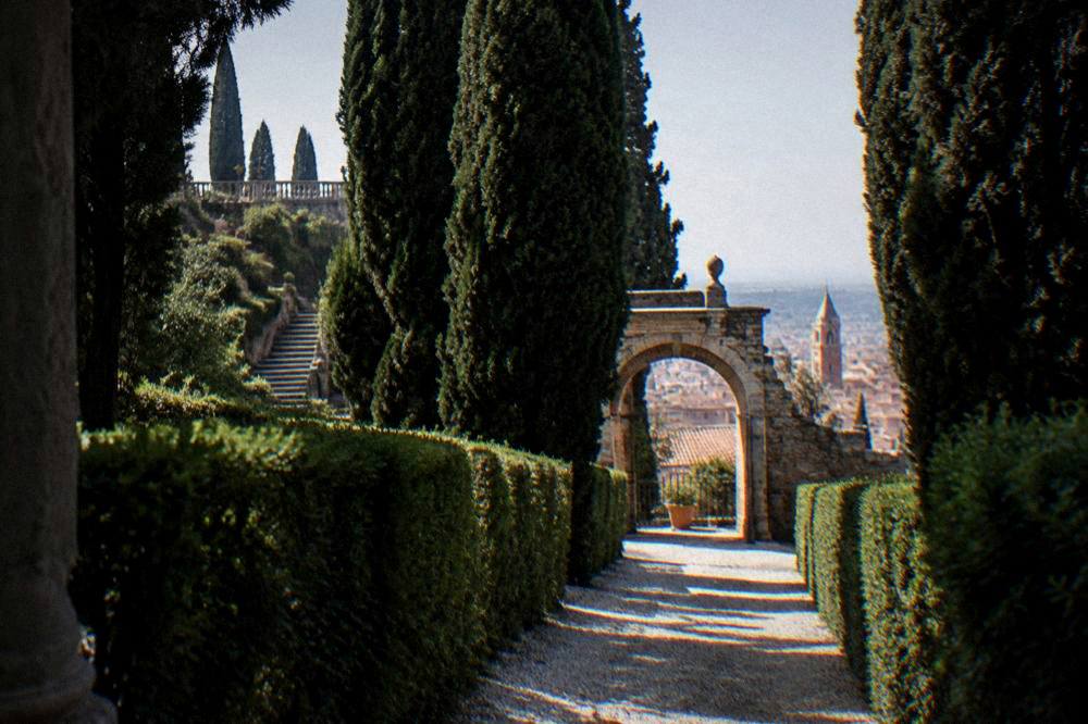 A shaded gravel alley between trimmed hedges leading toward an arched garden gate