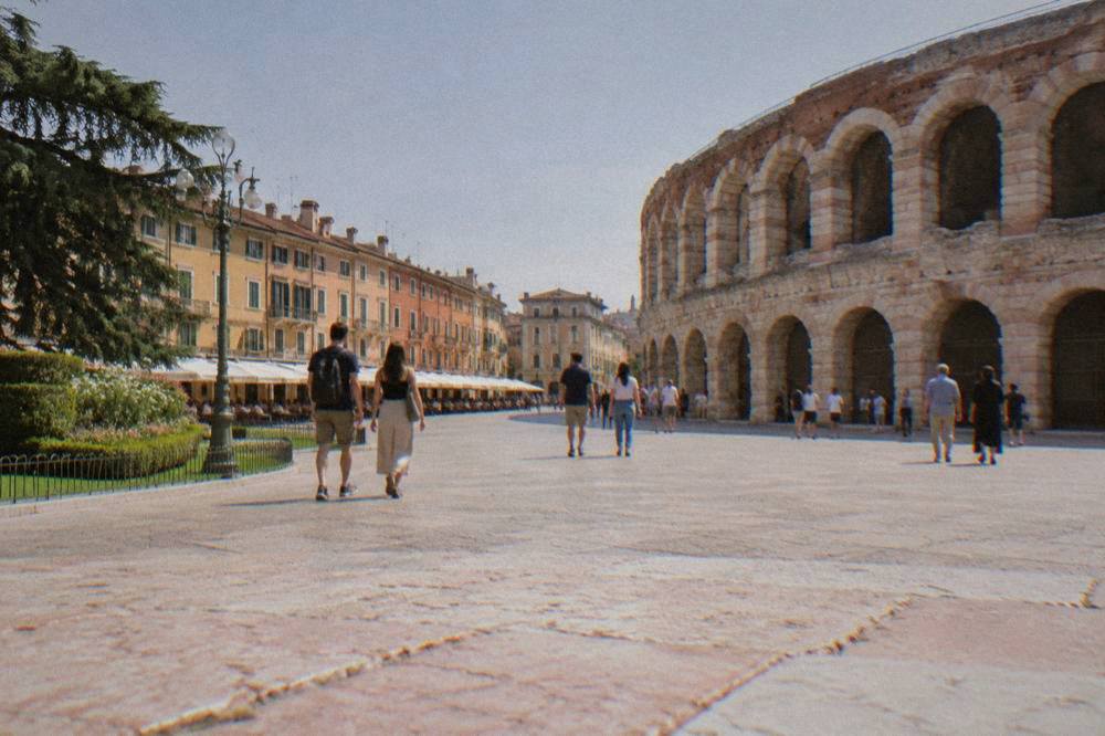 People walking across a broad city square toward the curved stone facade of the Verona Arena in morning light