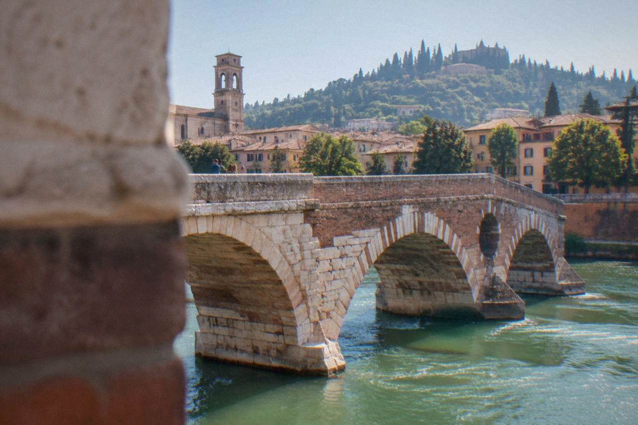 A stone Roman bridge with arches crossing a green river through a historic city