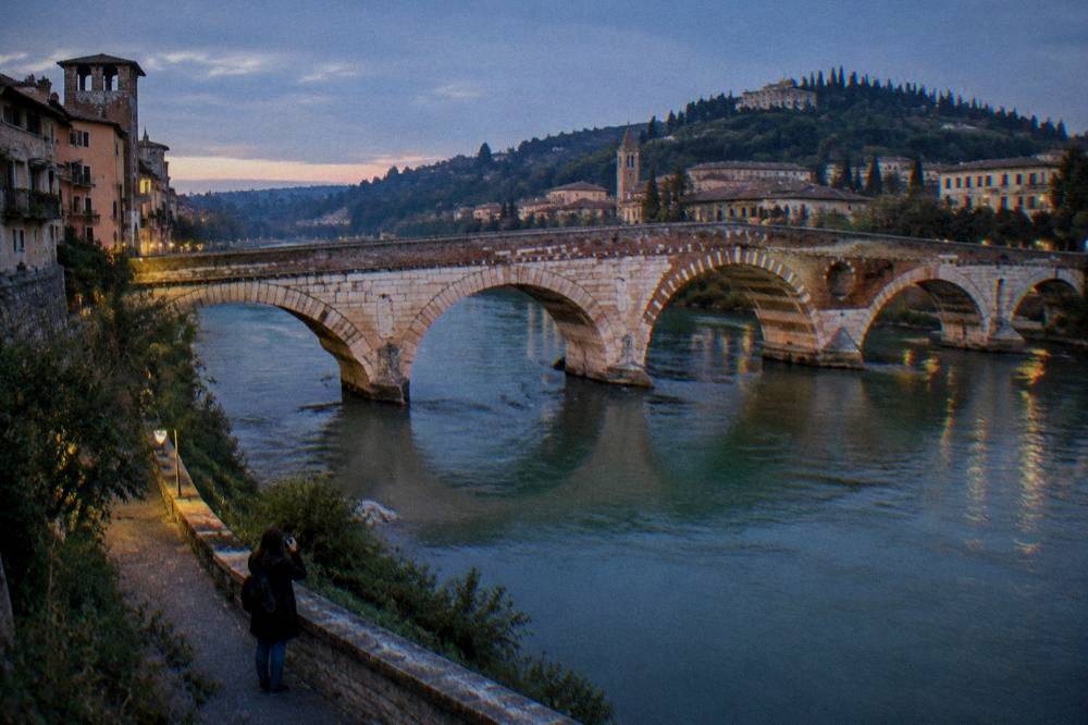 A photographer standing on a riverside path below an old stone bridge at sunset