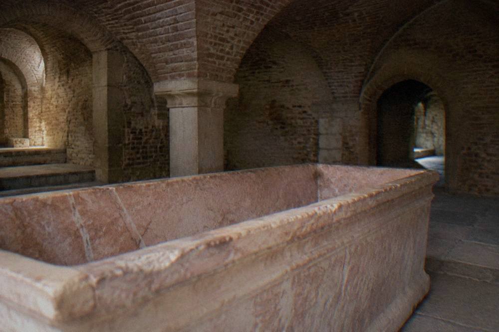 An empty open stone sarcophagus resting on the floor of a dim brick-vaulted crypt