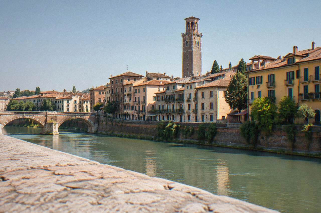 A tall medieval stone tower rising above the red rooftops of central Verona under a clear sky