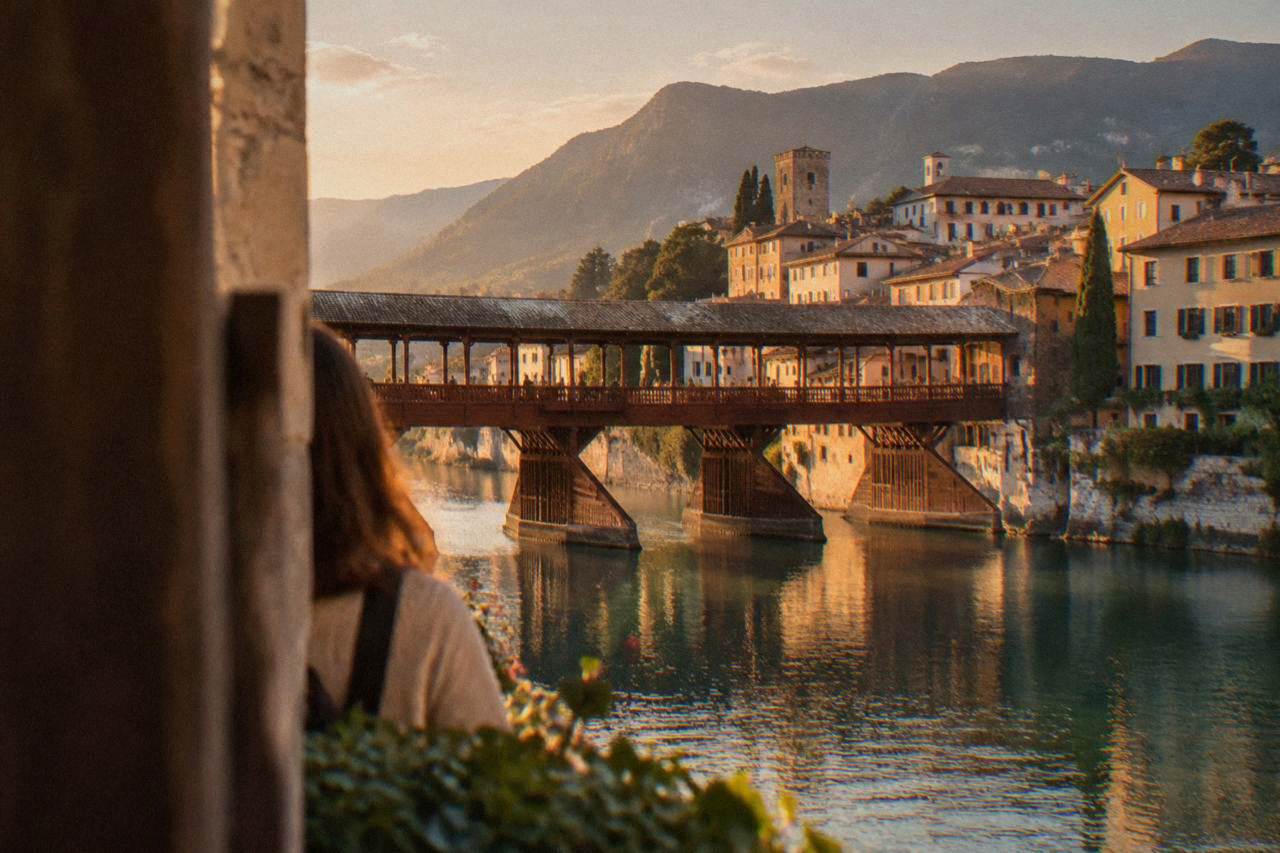 A covered wooden bridge spanning a mountain river with a town of tiled roofs behind it