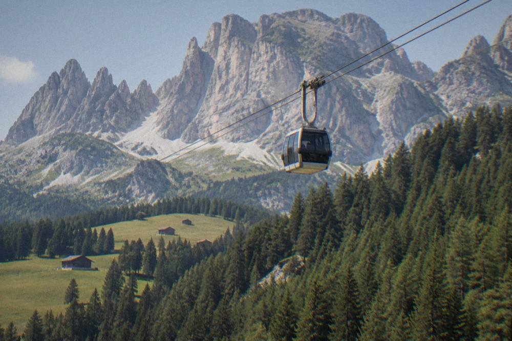 A cable car cabin rising above wooded slopes toward a rocky mountain ridge