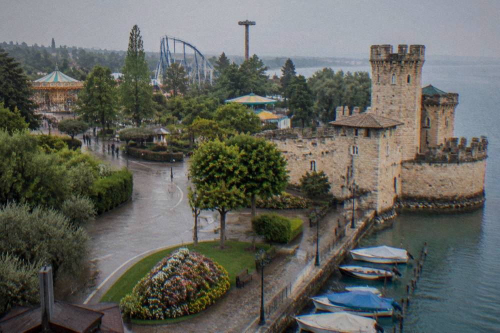 A stone castle tower above a lake promenade with boats moored alongside