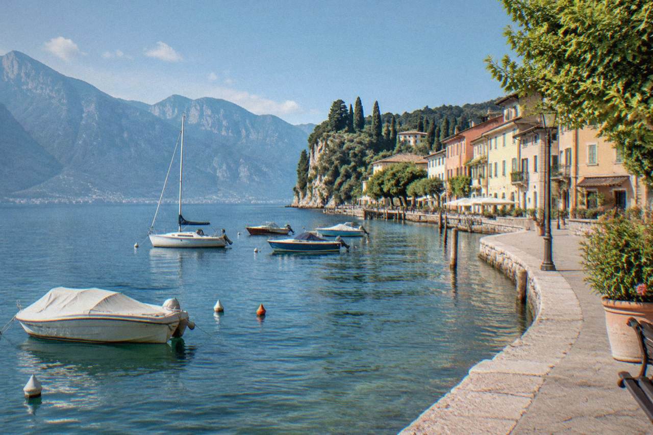 The shoreline of a Lake Garda town with boats moored in clear water and mountains rising behind