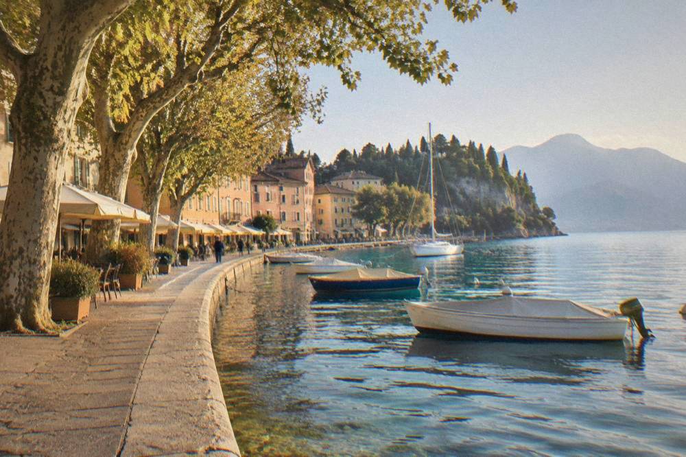 A lakeside promenade with plane trees, cafés and moored boats on a calm morning