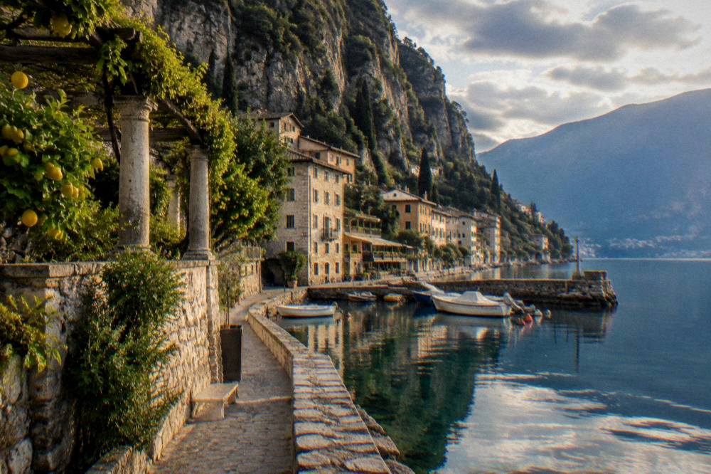 A quiet lakeside promenade with stone buildings and mountains reflected in still water at dusk