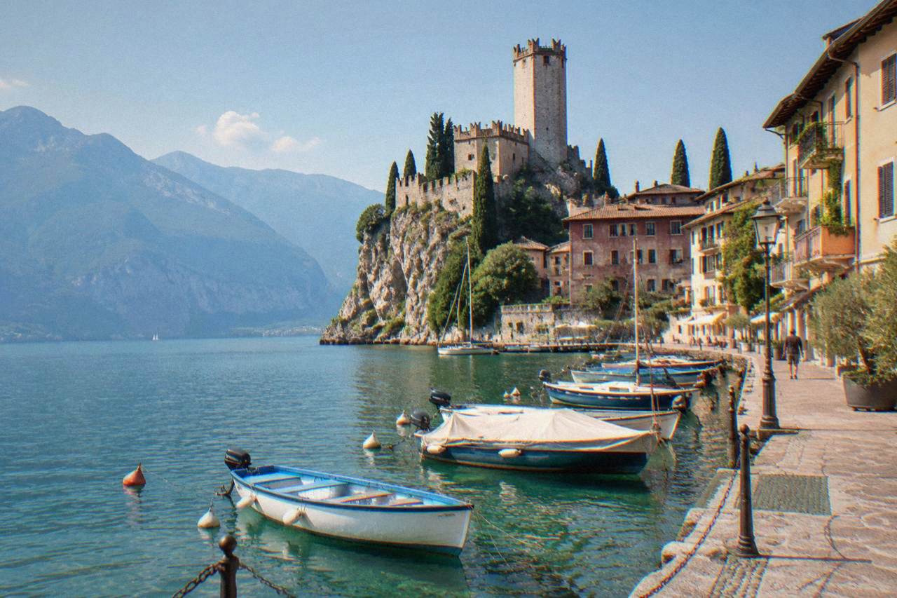 A lakeside town with a stone castle tower and boats moored along a harbour beneath steep mountains