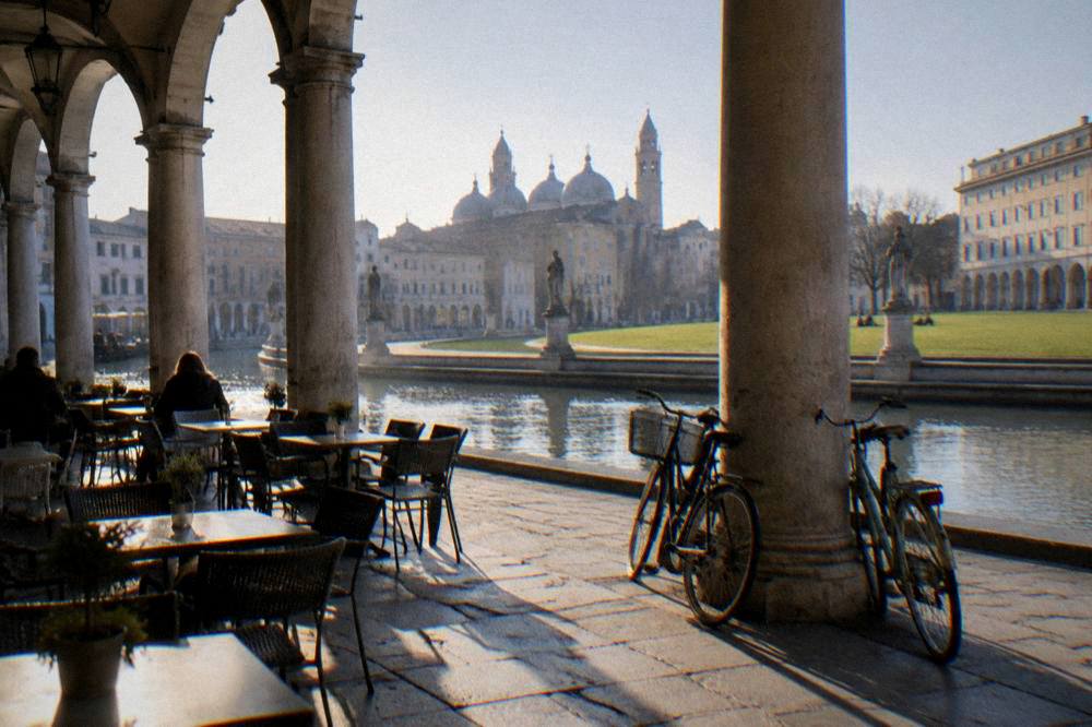 Sunlit cafe tables under an arcade in an Italian square with bicycles leaning against a column