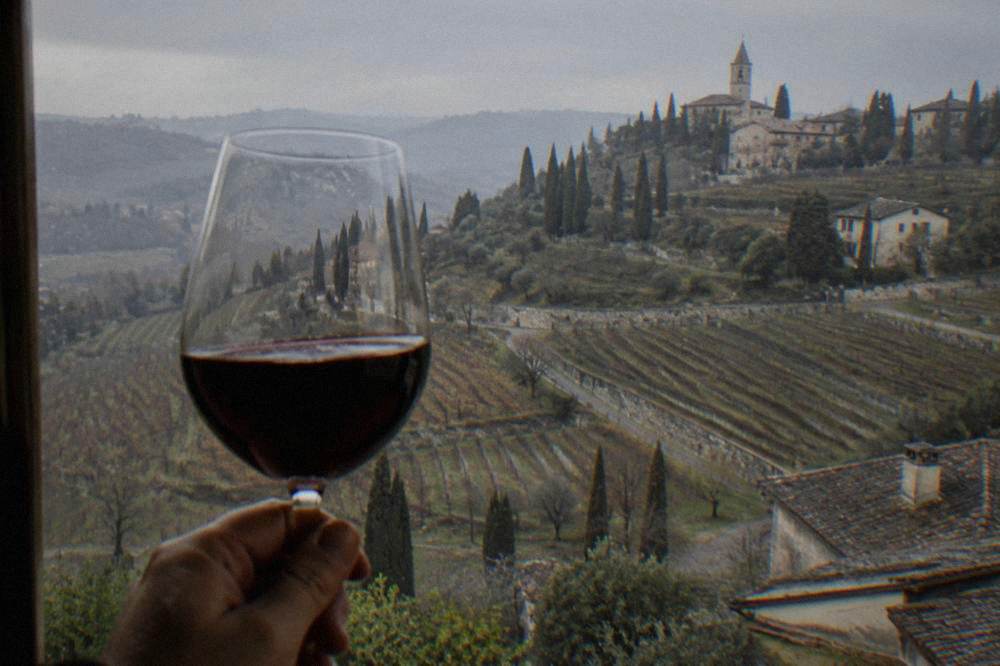A large glass of dark red wine held up against a window in low light