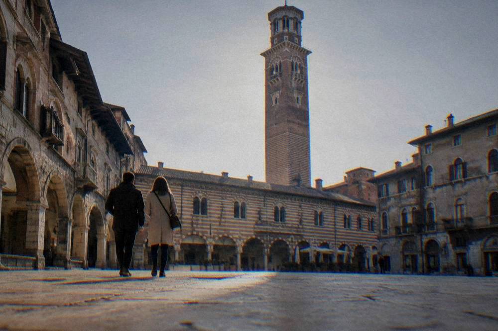 A couple walking across a Verona square with the Lamberti tower rising above the rooftops