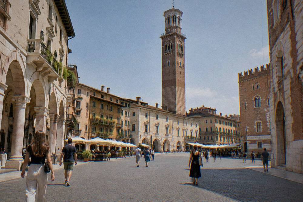 Pedestrians crossing a cobbled square in Verona with the Torre dei Lamberti rising above the rooftops