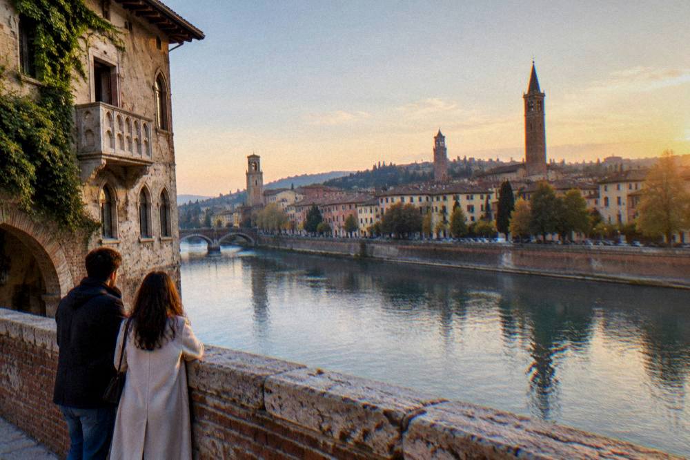Two people standing on an old stone bridge over a river at dusk with a city skyline behind