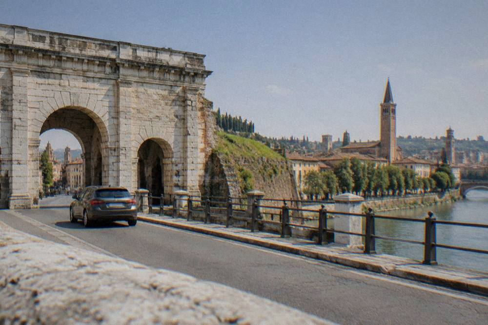 A car crossing a bridge over the Adige river with Verona's old town behind