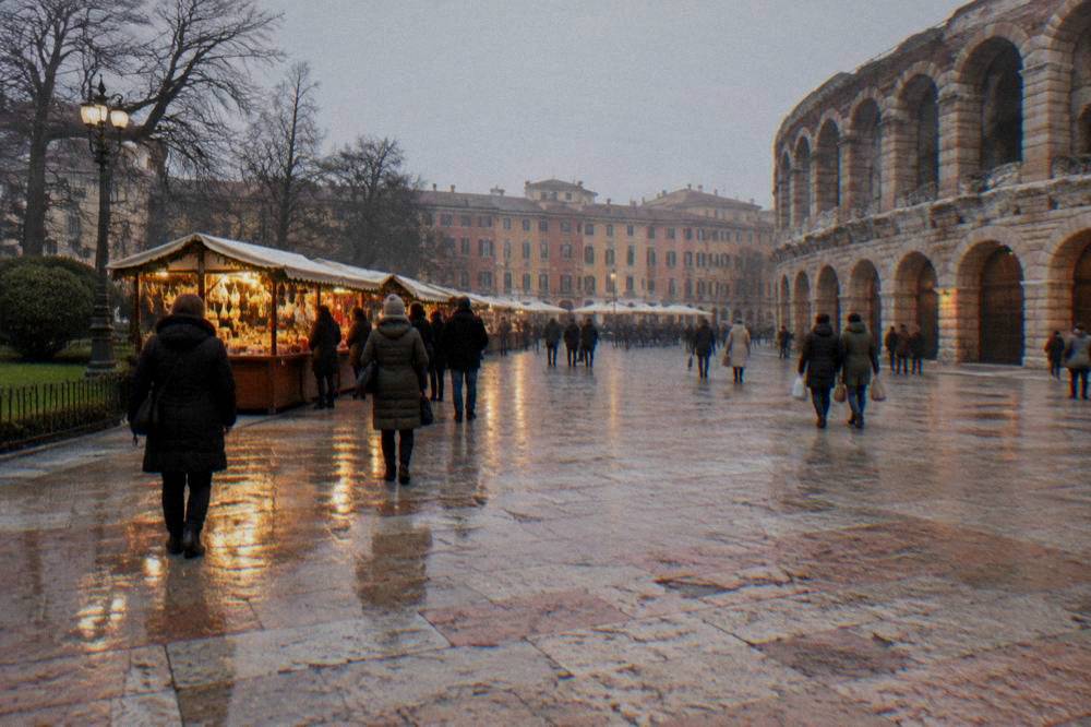Shoppers in warm coats walking past lit market stalls on a wet stone-paved square at night