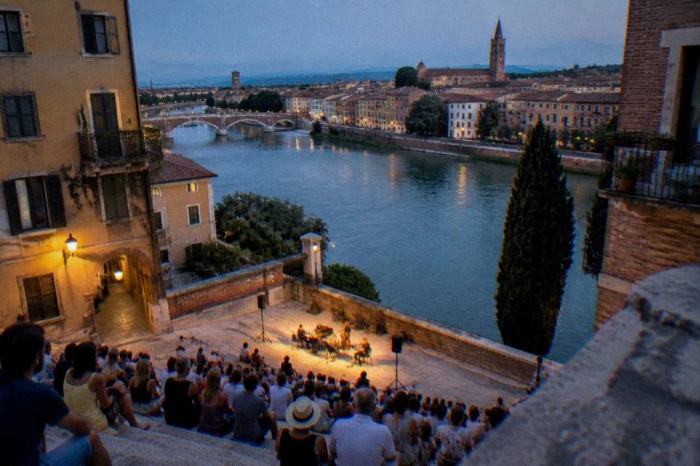 People in summer clothes seated on stone steps under evening lights at an open-air performance