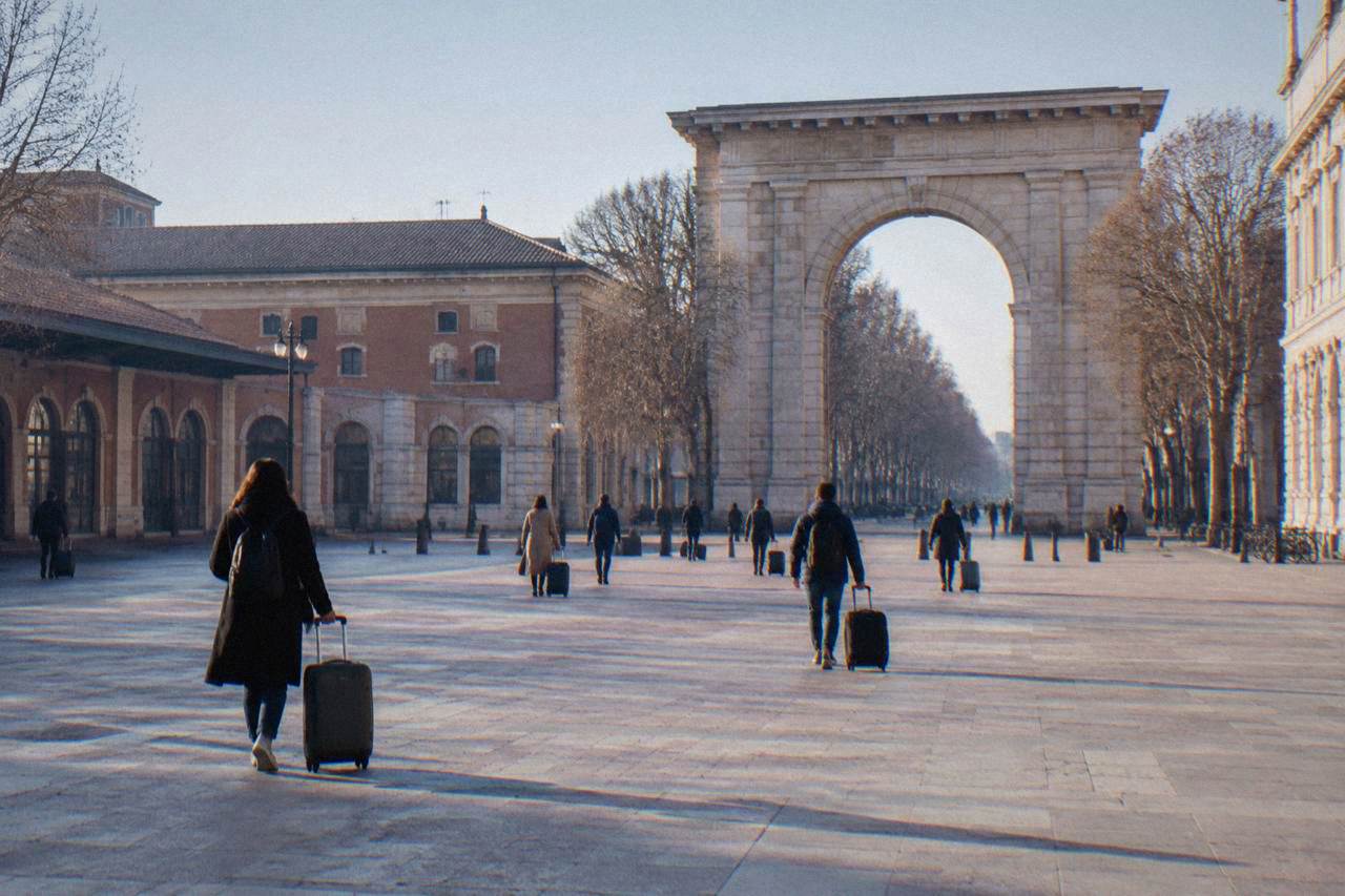 Travellers with suitcases walking across the wide open square in front of Verona Porta Nuova railway station on a bright morning