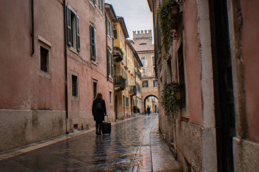A narrow cobbled lane in Verona's old town with rose-pink buildings and a person pulling a small wheeled suitcase