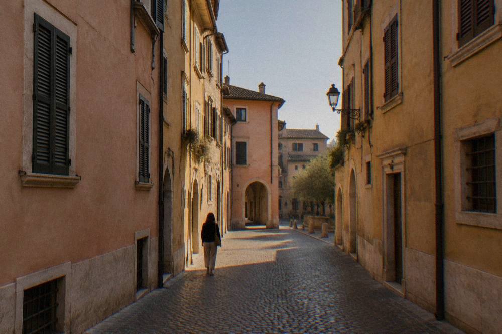 A narrow cobbled lane in central Verona with pastel walls, closed shutters and a single pedestrian walking through afternoon shade