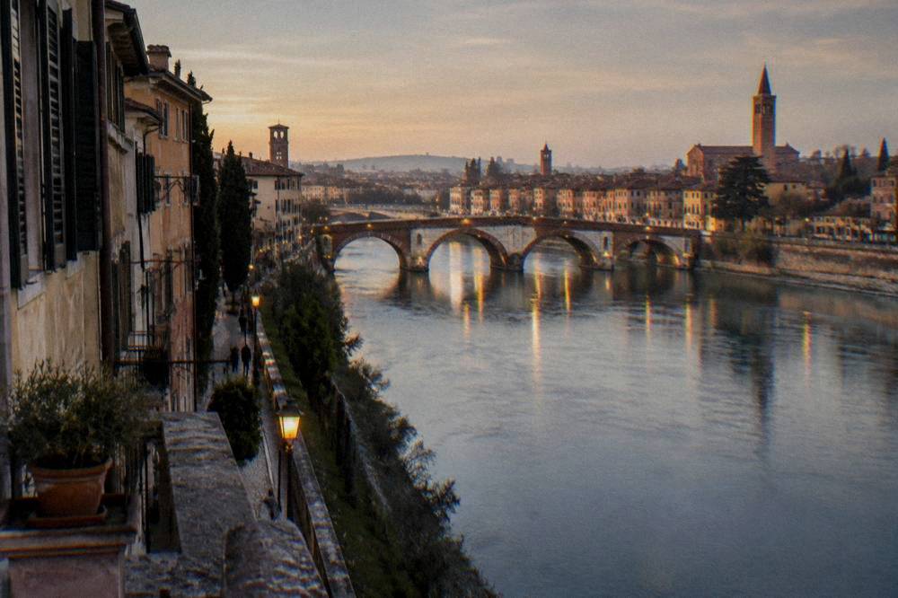 The Adige river in Verona at dusk with lit bridge arches reflecting on the water and the city skyline beyond