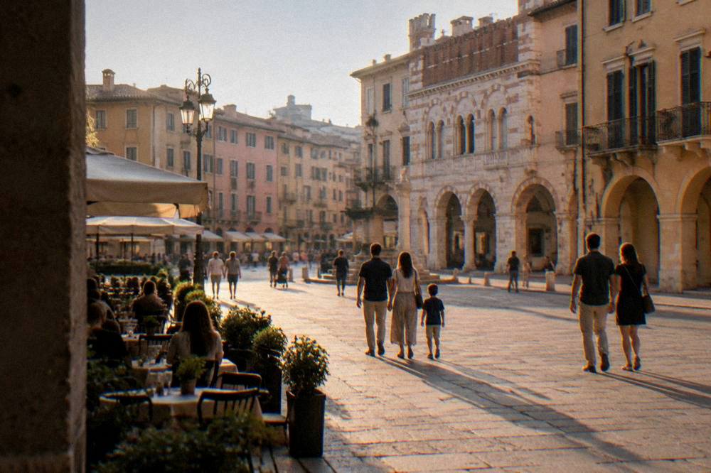 Families and couples strolling across a lit square in Verona in the evening with café tables along the edge