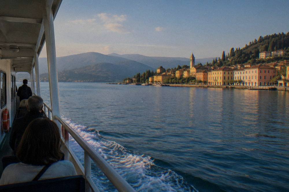 A ferry boat crossing blue lake water toward a lakeside town with hills behind