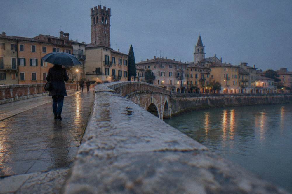 A visitor with an umbrella crossing an old stone bridge in Verona in light rain
