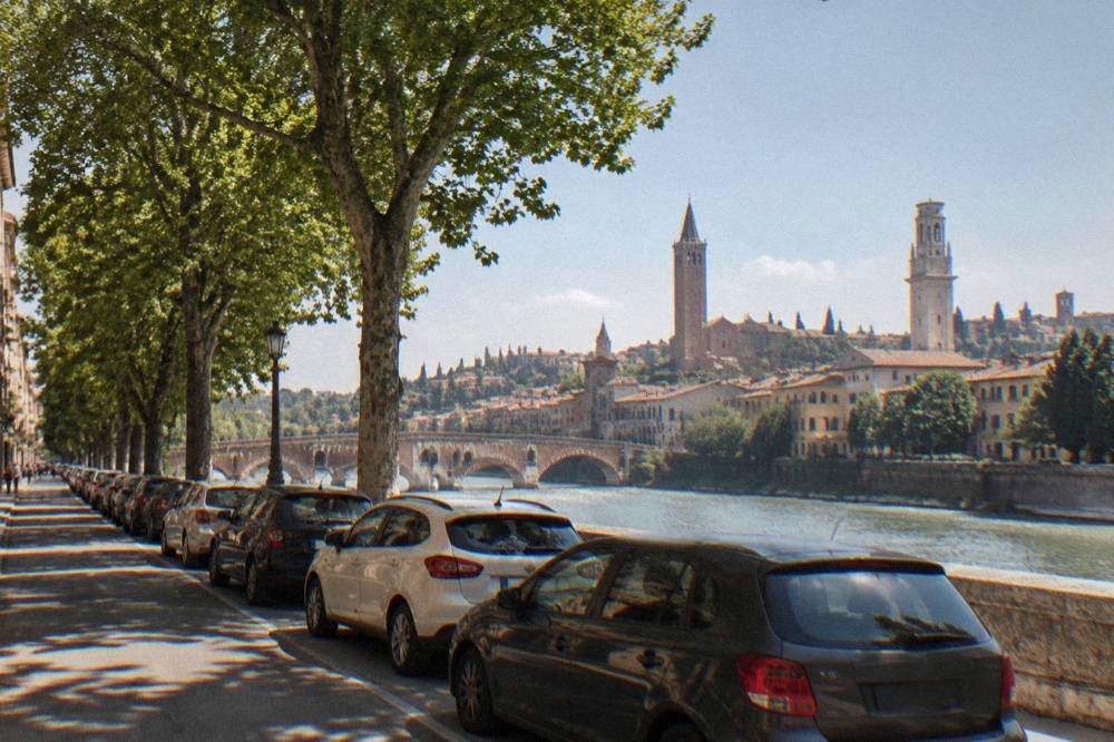 Rows of parked cars on a tree-lined street outside Verona's historic centre with the old city skyline behind