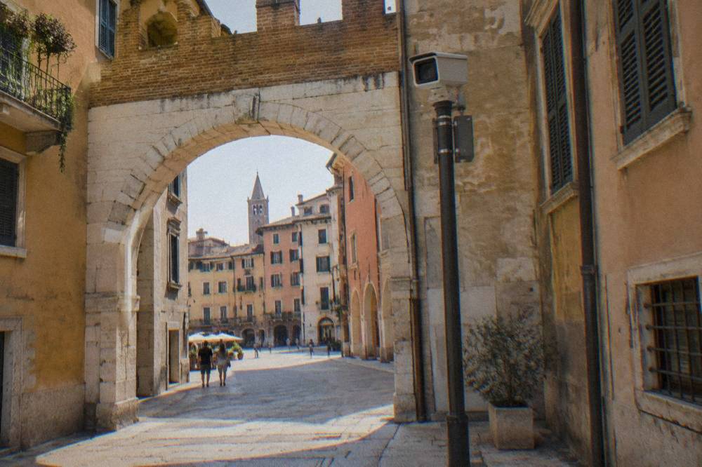 A traffic enforcement camera on a pole above a historic archway into a restricted street in Verona