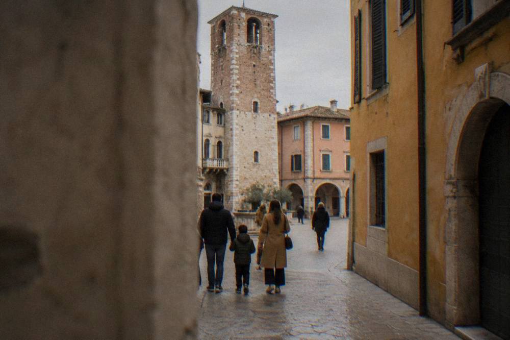 Parents and children crossing a narrow old street in Verona beside a warm stone tower